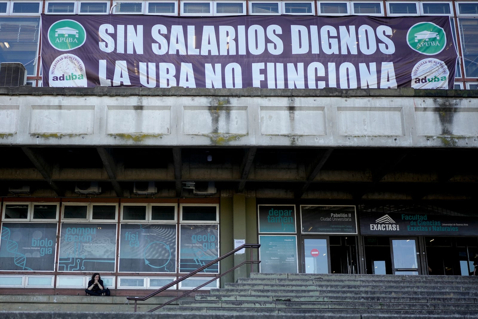 A student sits on the steps of the University of Buenos Aires (UBA) under a banner that reads in Spanish, "Without decent wages, UBA doesn't work," protesting President Javier Milei's veto of a law to increase funding for public universities in Buenos Aires, Argentina, Wednesday, Oct. 16, 2024. (AP Photo/Natacha Pisarenko)