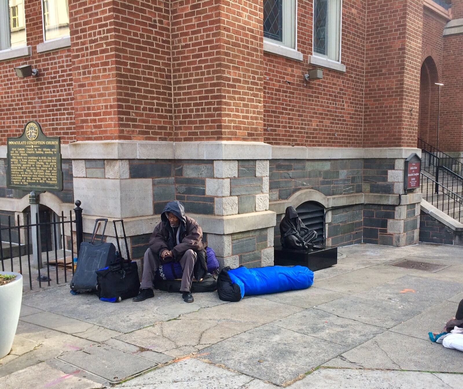 As evening dawns, homeless men gather to enter the Central Night Shelter run by the Catholic Shrine of the Immaculate Conception (behind the man) and the neighboring Central Presbyterian Church. The homeless man pictured said he knew Ricky Bradfield, who recently died, but never talked with him. “What would I talk to him about?” said the man who wouldn’t give his name but eerily resembled the Shrine statue on the right. 