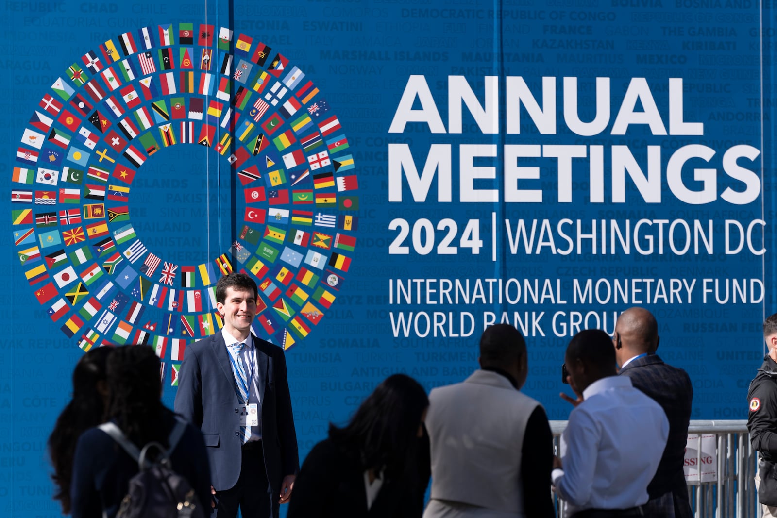 An attendee poses for a photograph in front of a sign advertising the annual meetings of the International Monetary Fund (IMF) and the World Bank Group (WBG), which started in Washington on Monday, Oct. 21, 2024. (AP Photo/Ben Curtis)