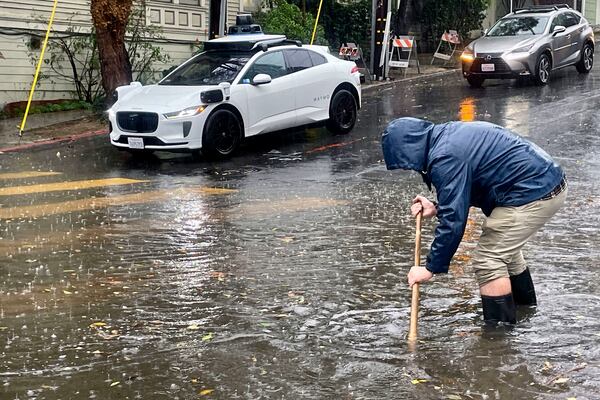 A Mission District resident attempts to unclog a few storm drains on a flooded street as a driverless Waymo taxi passes in the background, Friday, Nov. 22, 2024, in San Francisco. (AP Photo/Haven Daley)