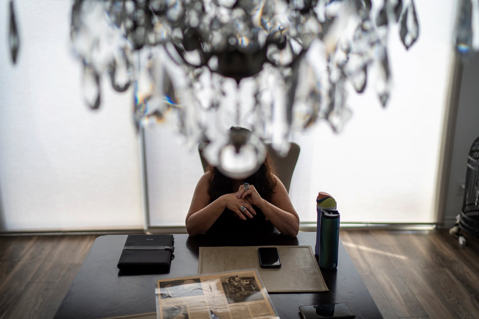 Buttons sits obstructed by a chandelier with a laminated newspaper clipping of her as a child and her adopted parents in Wisconsin, as she visits a friend, Monday, June 24, 2024, in Henderson, Nev. Buttons fears being deported back to Iran, so carries around the newspaper clipping, stacks of adoption files and court records as proof she's supposed to be here. (AP Photo/David Goldman)