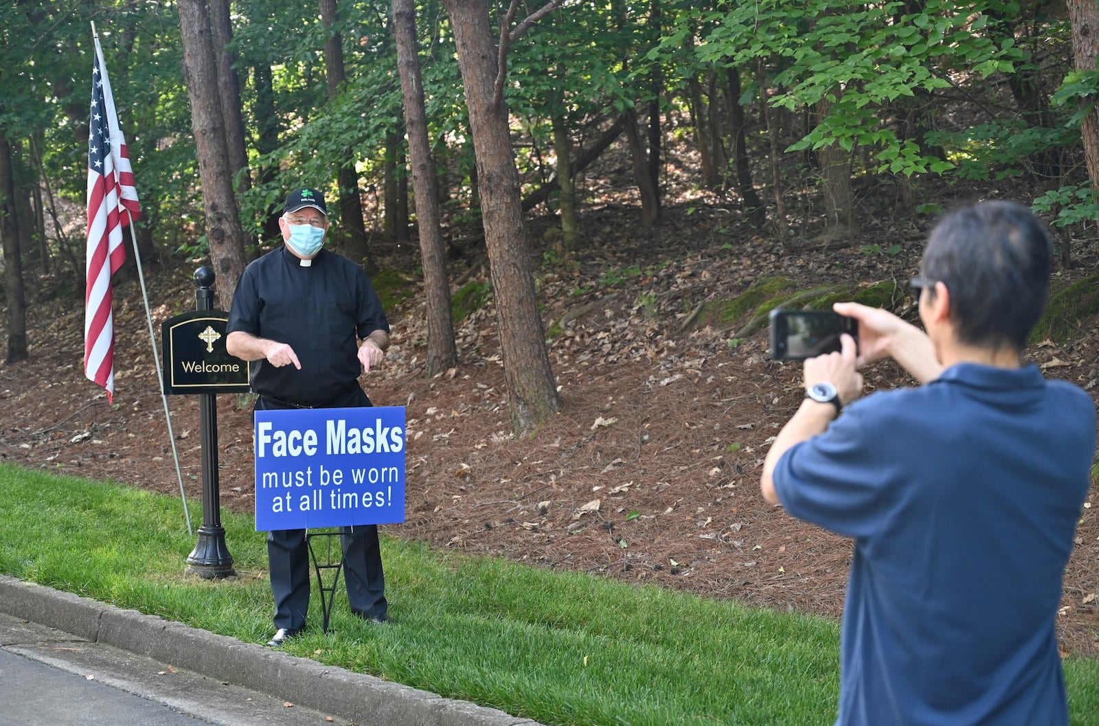 Father Paul Flood poses in front of a sign as Khanh Mai, facility manager, takes a photograph using a smartphone on Saturday, May 23, 2020, at the entrance of St. Benedict Catholic Church. Daily Mass is expected to resume Monday. (Photo: Hyosub Shin / Hyosub.Shin@ajc.com)