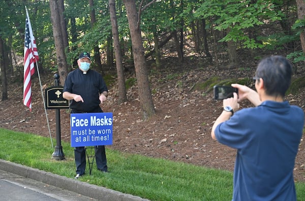 Father Paul Flood poses in front of a sign as Khanh Mai, facility manager, takes a photograph using a smartphone on Saturday, May 23, 2020, at the entrance of St. Benedict Catholic Church. Daily Mass is expected to resume Monday. (Photo: Hyosub Shin / Hyosub.Shin@ajc.com)