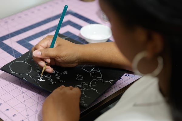 Bria Bowen, who has a degree in graphic design, works on a custom graduation hat for a client in her home office in Kennesaw on Wednesday, April 19, 2023. (Natrice Miller/natrice.miller@ajc.com)