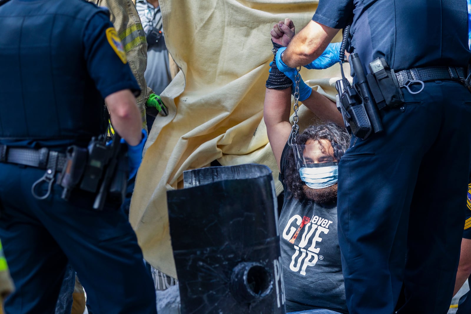 Tallahassee police handle protester Jordan Mazurek, 28, who cemented his hands in two 55-gallon plastic drums filled with concrete in the driveway of the Governor's Mansion, Friday, April 17, 2020, in Tallahassee, Fla. Mazurek is protesting how Gov. Ron DeSantis is handling coronavirus in state prisons. (Alicia Devine/Tallahassee Democrat via AP)