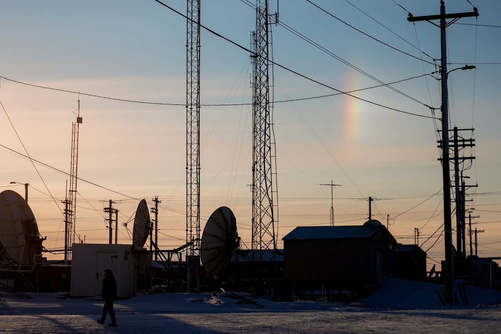 A sundog forms a vertical rainbow as a villager walks by transmission dishes in Kaktovik, Alaska, Tuesday, Oct. 15, 2024. (AP Photo/Lindsey Wasson)