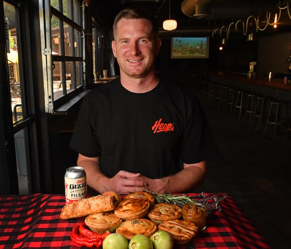 230818 Atlanta, Ga: Heaps Atlanta owner, Jake Harvey, with a few of the products he offers, including (in no particular order): Pork & Sage Salsa Roll, Carnitas Al Pastor Pie, Chana Masala Pie, Lamb & Vegetable Pie, Chicken Pot Pie, Green Tomato Pie, Beef & Cheddar Pie, photographed with the award winning Heaps Gizza - New Zealand Style Pilsner. The AJC visits chef Jake Harvey at his business, Heaps Atlanta, in the Boggs Social building at 1310 White Street SW 30310 in Atlanta, Ga. PHOTO FOR FOOD COVER STORY ON NEW ZEALAND MEAT PIES AAJC090623MEATPIES (CHRIS HUNT FOR THE ATLANTA JOURNAL-CONSTITUTION)