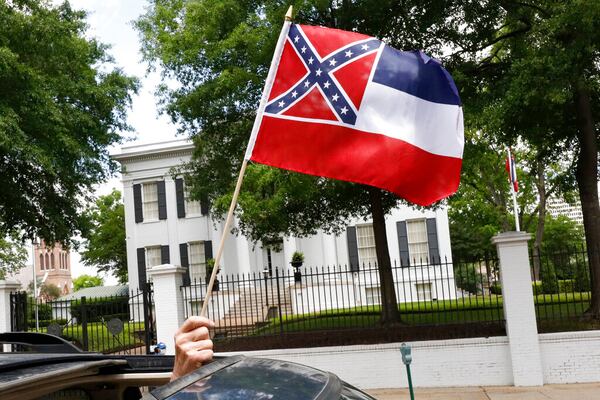 In this April 25, 2020 photograph, a small Mississippi state flag is held by a participant during a drive-by "re-open Mississippi" protest past the Governor's Mansion, in the background, in Jackson, Miss. This current flag has in the canton portion of the banner the design of the Civil War-era Confederate battle flag, that has been the center of a long-simmering debate about its removal or replacement. (AP Photo/Rogelio V. Solis)