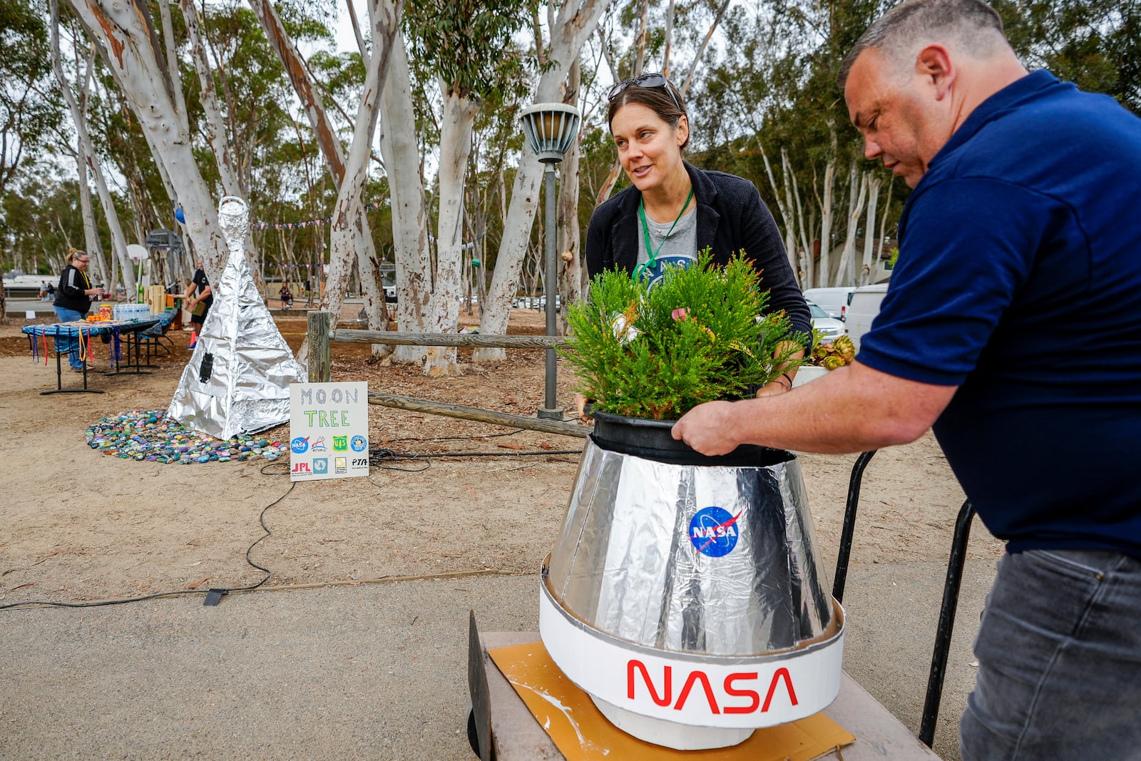 Santiago parent volunteer Stacie Aguesse, left, delivers a small Giant Sequoia tree from NASA's Artemis I Mission's tree seeds that traveled around the moon twice, as NASA scientists, JPL engineers, US Forest Service representatives, and teachers join Santiago STEAM Magnet Elementary School students at a ceremony to plant it after the school was honored in the spring of 2024 to become NASA Moon Tree Stewards in Lake Forest, Calif., on Monday, Oct. 14, 2024. (AP Photo/Damian Dovarganes)