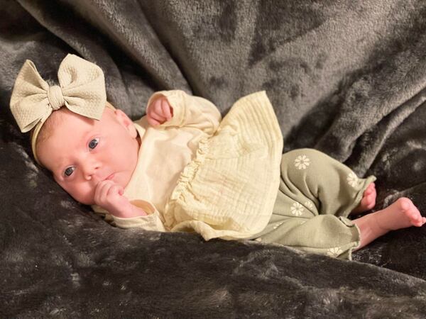 Baby Eleanor Ruth McGee is laying on her side side on a soft, brown blanket. She has a large pale yellow bow on her head, and is wearing a pale yellow long-sleeve shirt of the same color and matching pants with a pale yellow flower pattern.