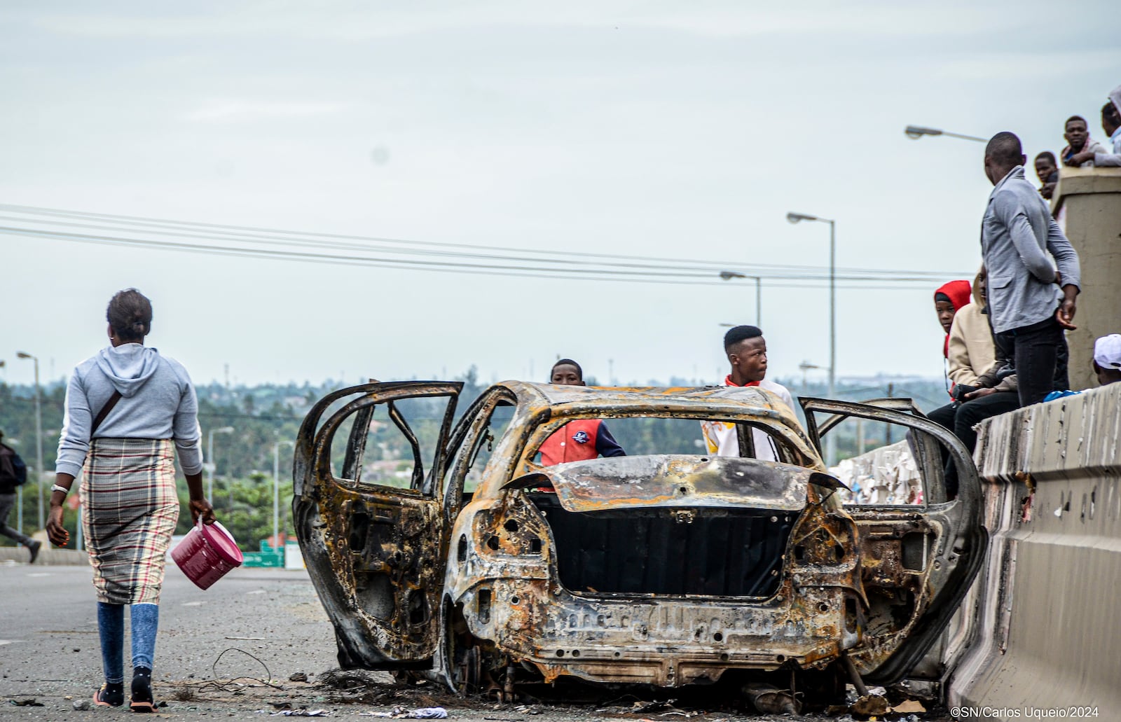 Pedestrians alongside burnt out vehicle Wednesday, Nov. 6, 2024 in Mozambique's capital Maputo, in protests that have engulfed the country after the opposition rejected the results of the country's polls which saw the Frelimo party extend its 58-year rule. (AP Photo/Carlos Uqueio)