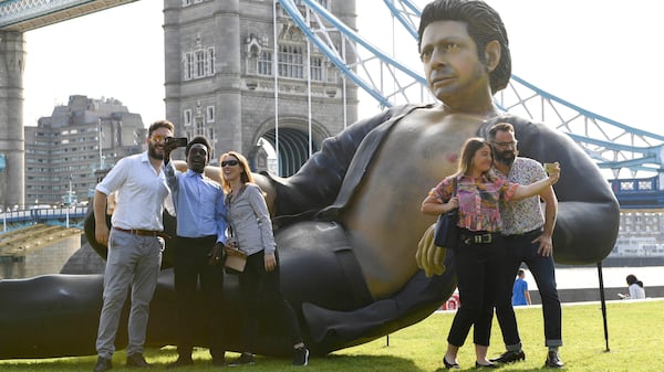 People take photos by a 25ft statue of actor Jeff Goldblum in a pose from a scene in the first Jurassic Park movie, which has been created by a TV channel to celebrate the film's 25th birthday, at Potters Fields Park, London, Wednesday July 18, 2018. Tower Bridge in the background.