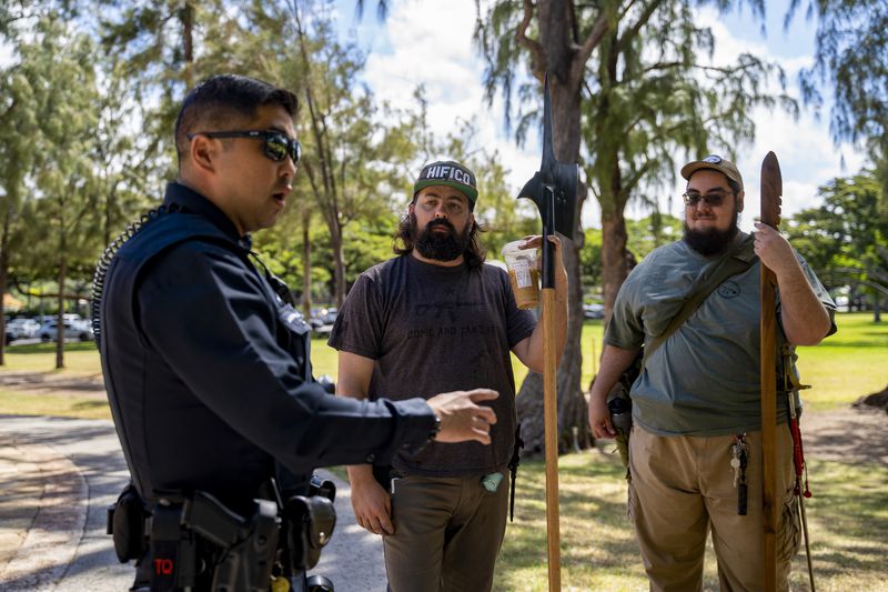 Andrew Roberts, center, director of the Hawaii Firearms Coalition, holds a halberd while talking to a Honolulu police officer at Kapiolani Park on Saturday, June 22, 2024, in Honolulu, Hawaii. (AP Photo/Mengshin Lin)