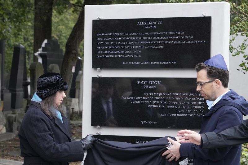 Yuval Danzig, right, the son of a Polish-Israeli man kidnapped and killed by Hamas, helps to unveil a plaque honoring his father, Alex Dancyg, in the Jewish cemetery on the one-year anniversary of the Hamas attack, in Warsaw, Poland, on Monday, Oct. 7, 2024. (AP Photo/Czarek Sokolowski)