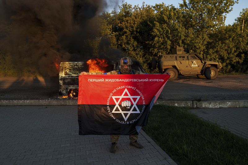 A Ukrainian serviceman holds a flag for his fallen comrade in front of a burned vehicle near Russian-Ukrainian border, Sumy region, Ukraine, Wednesday, Aug. 14, 2024. (AP Photo/Evgeniy Maloletka)