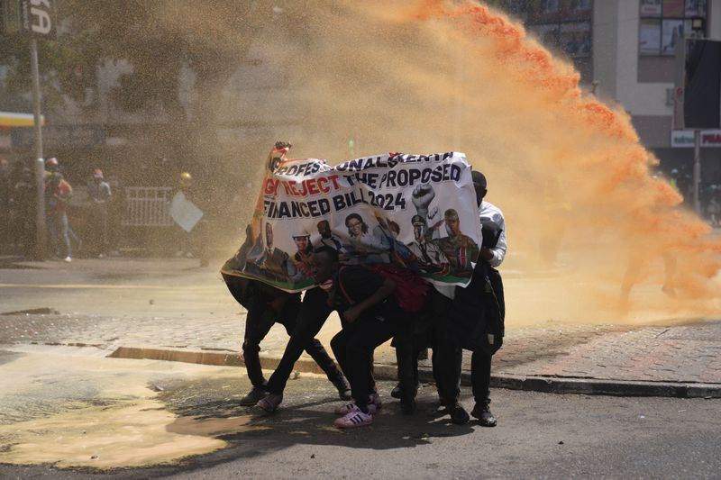 Protesters hide behind a banner as Kenya police spray water cannon at them during a protest over proposed tax hikes in a finance bill in downtown Nairobi, Kenya Tuesday, June 25, 2024. (AP Photo/Brian Inganga)