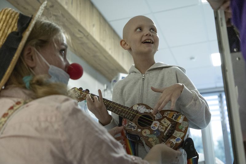 Tetiana Nosova, who goes by the clown name of Zhuzha, a volunteer from the "Bureau of Smiles and Support" watches as Kira Vertetska, 8, plays a ukulele at Okhmatdyt children's hospital in Kyiv, Ukraine, Thursday Sept. 19, 2024. (AP Photo/Anton Shtuka)