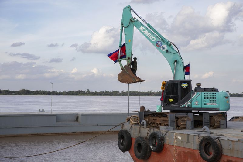 A worker raises a Cambodian flag with the help of an excavator on a barge by the mouth of a canal, at Prek Takoe village, Phnom Penh, Cambodia, Tuesday, July 30, 2024. (AP Photo/Anton L. Delgado)