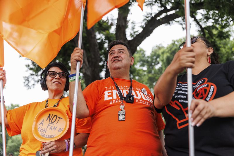 Robb Elementary shooting survivors Amy Franco, left, Arnulfo "Arnie" Reyes, center, stand with other survivors and community members at the town square on Friday morning, May 24, 2024, in Uvalde, Texas. The former Robb Elementary School educators waved orange flags signifying gun violence awareness to commemorate the 21 victims of the shooting – 19 fourth-graders and two teachers – who died two years ago Friday. (Sam Owens/The San Antonio Express-News via AP)